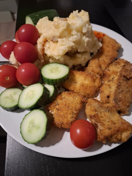 Panerad fisk med potatismos, tomater och gurkskivor på en tallrik.