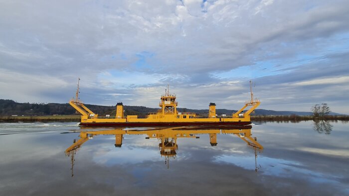 Bilfärja Linea på Göta älv, på väg från Stockholm till Vänersborg för att ersätta Dalbobron, speglas i vattnet under en molnig himmel.