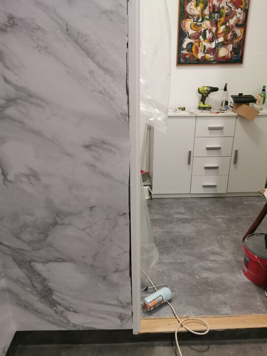 Badrum under renovering med grå marmorvägg och golvmaterial, slarvigt installerad matta vid dörrkarm och tröskel, verktyg synliga i rummet.