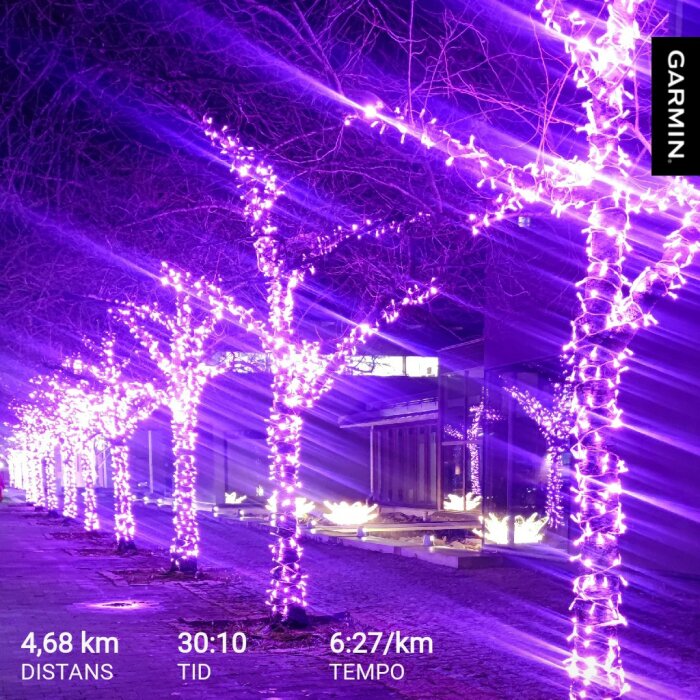 Rad av träd dekorerade med lila ljusslingor på natten, med en byggnad i bakgrunden och text om distans och tid för löpning.