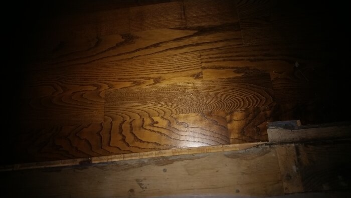 Trägolv i borstad ask med mörka ådringar och glans, potentiellt från Barlinek, monterat ovanpå en grund av rå trästruktur.