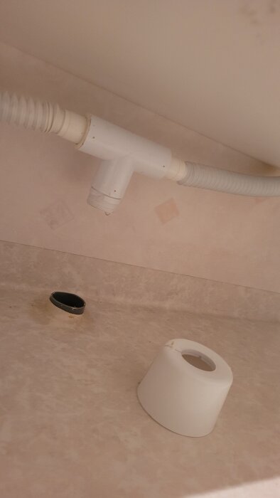 Avloppsrör under dubbelhandfat med en T-koppling och löst liggande vit plastdel bredvid.