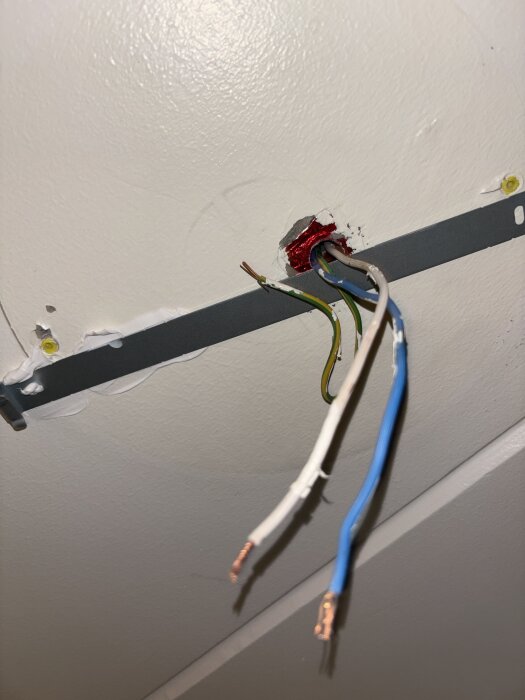 Elektriska kablar som sticker ut från ett vägguttag, med en vit, blå och gul-grön ledning synlig mot en vit vägg.