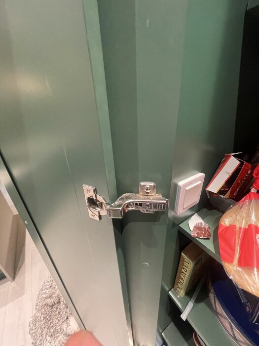 Grön skåpsram monterad på täcksidor till kyl och frys med synlig gångjärnsmekanism, omgiven av hyllor med matvaror.