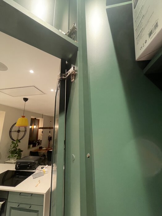 Grön skåpsram med gångjärn, fäst mellan täcksidor för kyl och frys i ett kök. Vitt köksbänk och köksutrustning syns i bakgrunden.