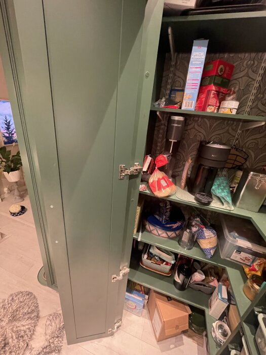 Grön köksskåpsram fäst på täcksidor, dörrar med gångjärn öppnas mot ett fullt mellanslag med förvaringshyllor fyllda med diverse föremål.