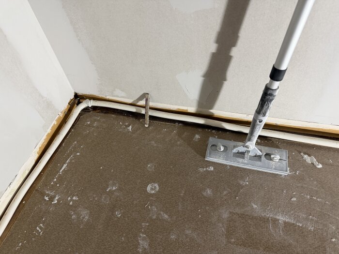 Hjørne af et rum, hvor gulvlister og rør er synlige. En moppe med hvidt skaft står i hjørnet, og gulvet har pletter af maling eller støv.