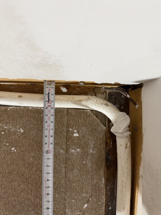 Foto af en væg ved hjørne med en hvid rørinstallation og en lineal, der måler afstanden fra væggen. Træpaneler og vægmateriale ses bagved.