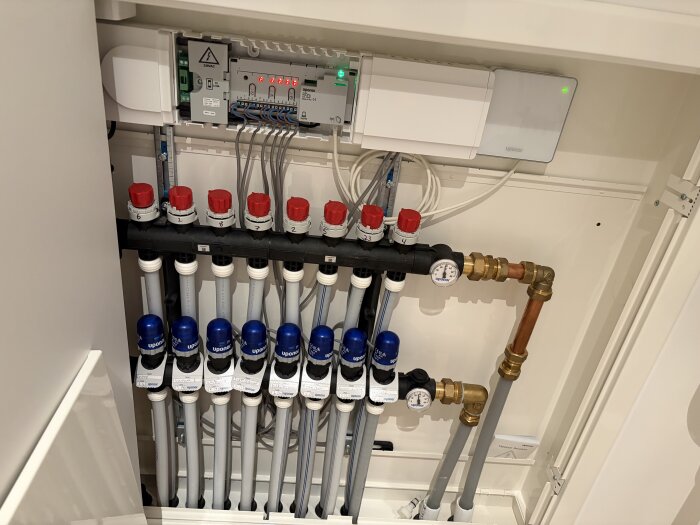 Hydroniskt golvvärmesystem med rör, ventiler och en kontrollenhet, sannolikt Uponor. Flera kopparrör och kabelanslutningar syns.