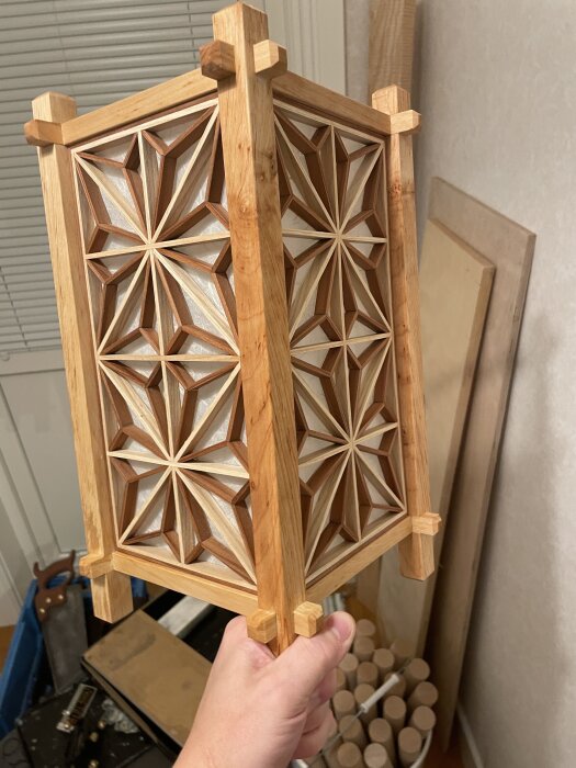 Trähantverk med intrikat geometriskt mönster, hållen i en hand framför fönster och trädetaljer i bakgrunden.