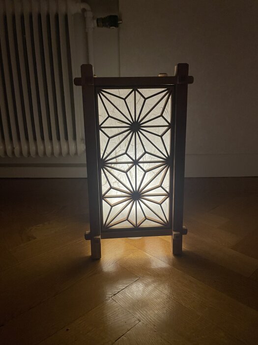 Lampa med geometriskt mönster och trädetaljer, placerad på trägolv vid en radiator.