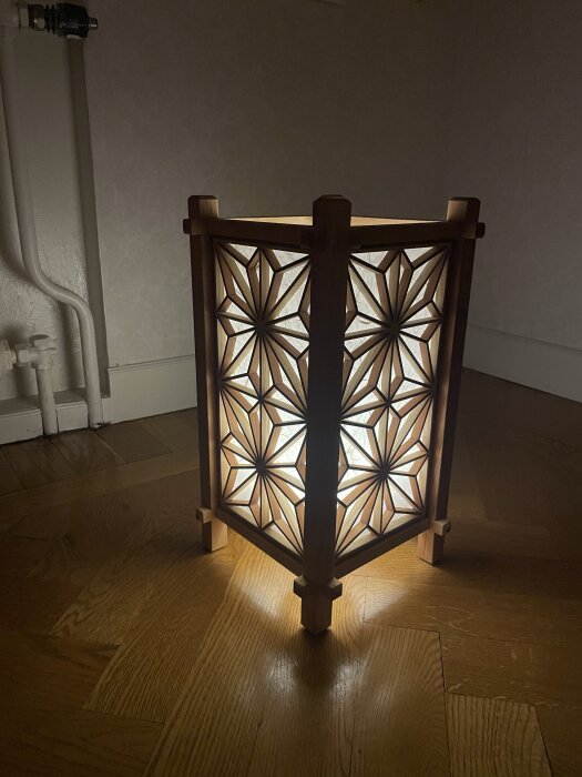 En vacker, tänd trälykta med geometriskt mönster står på ett trägolv i ett hörn av ett rum.