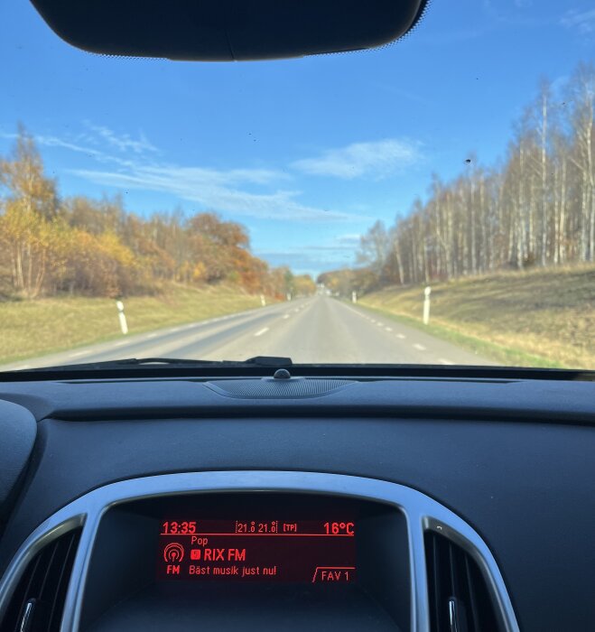 Bilinteriörsbild visar instrumentpanel med RIX FM på radion och utsikt över rak landsväg kantad av höstträd och blå himmel.