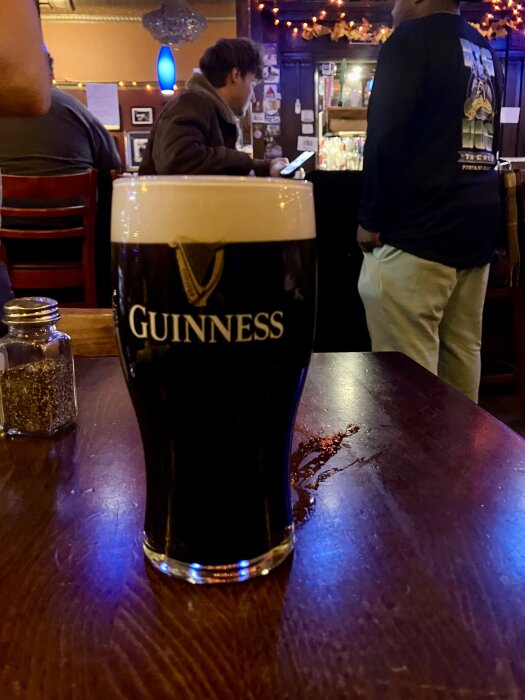 Ett glas Guinness-öl på ett träsbord i en mysig pubmiljö med människor i bakgrunden.