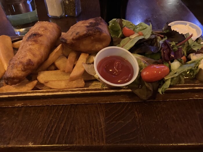Fish and chips med två stora fiskfiléer, pommes, ketchup, grönsallad och ett glas Guinness på träplanka.