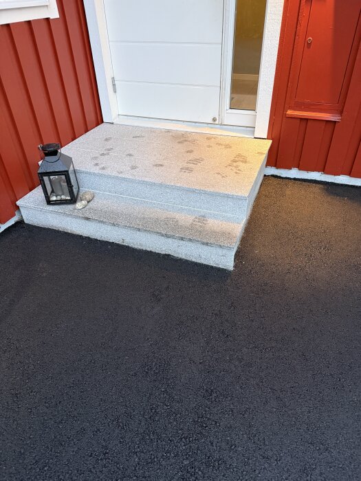 Nyasfalterad uppfart med betongtrappa och lanterna, framför en röd husvägg.