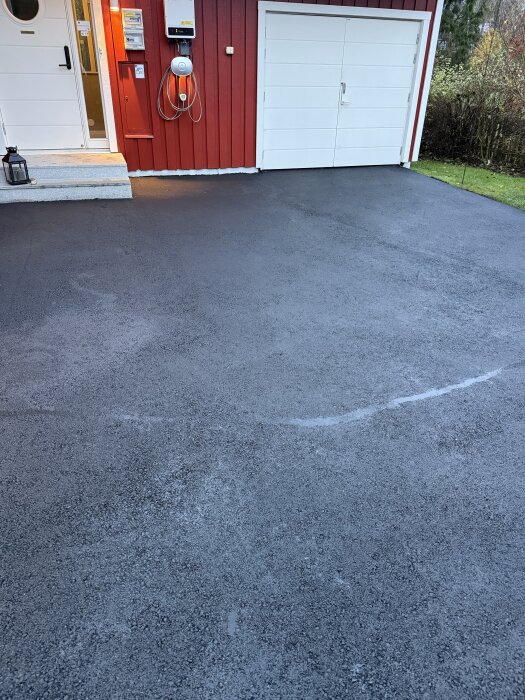 Nyasfalterad uppfart framför ett rött hus med vit dörr och garage.