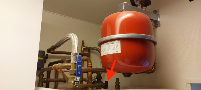 Rött expansionskärl med rörledning och blå ventil i närbild, röd pil pekar på justeringsventil under kärlet.