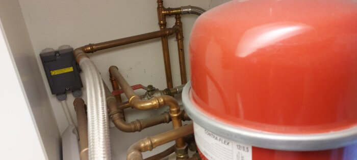 Expansionskärl med kopparrör, ventil med röd spak och en anslutningslåda på en vägg inomhus.