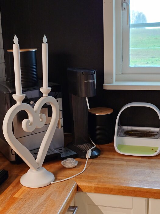 En köksbänk med en vit hjärtformad ljusstake, en kaffemaskin, och en Sodastream vid ett fönster.