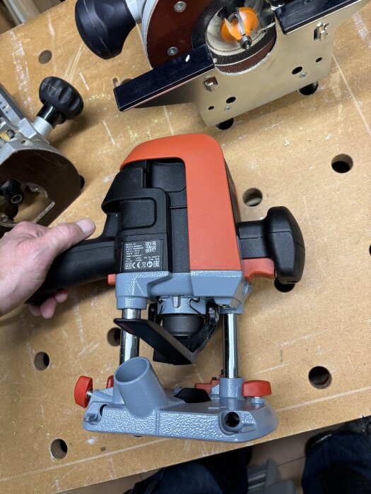 Hand håller i en mellanfräs på ett arbetsbord, omgiven av andra verktyg. Fräsen har en röd och grå färgsättning med uttag för dammutsugning.