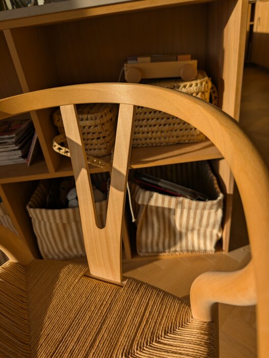 Trästol med y-formad rygg, lätt gulnad, står framför hylla med korgar, morrnarna ljus faller mjukt över ytan.