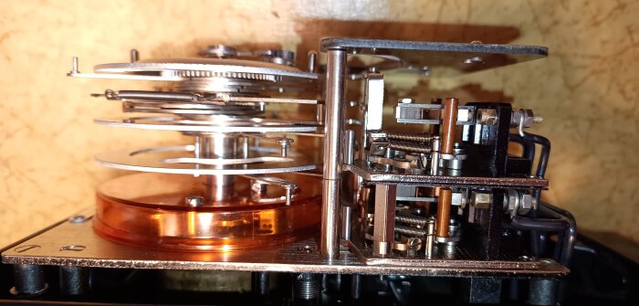 Avancerat mekaniskt verk av ett ovanligt tidur från Saia AG, visande finmekanik och kopparfärgade detaljer mot en träliknande bakgrund.