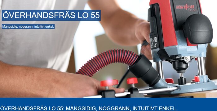 Hand håller i en Mafell LO 55 överhandsfräs som används för träbearbetning. Röd och blå design med flexibel sugslang.
