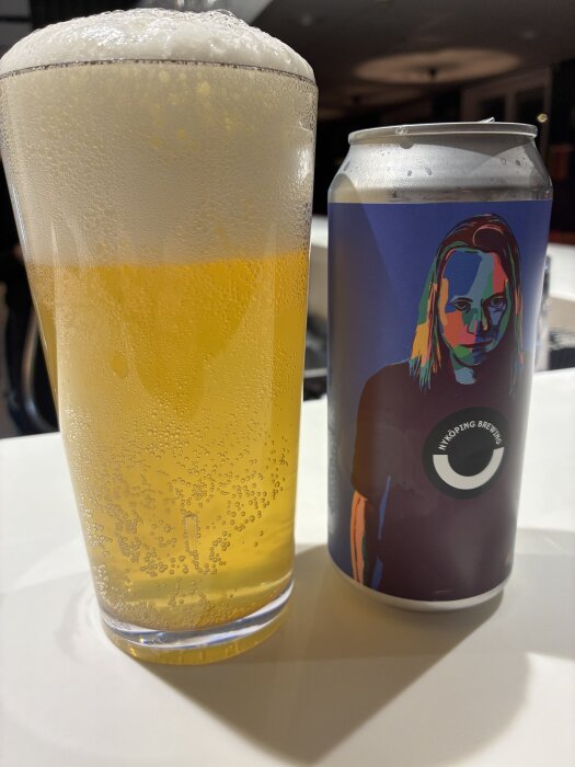 Ölglas med skummande pilsner bredvid en färgglad burk från Nyköping Brewery på ett bord.