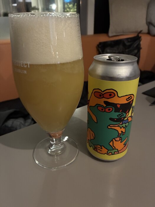 Ett glas hazy IPA med rikligt skum och en färgglad burk med tecknade figurer på ett bord.