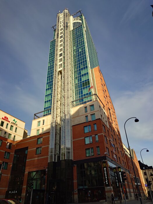 En hög byggnad med glasfasad mot en blå himmel. I bottenvåningen finns skyltar för McDonald's, ICA och andra butiker.