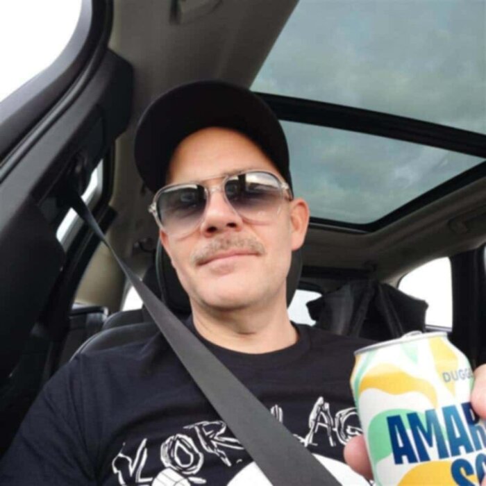 Person i bil med keps och solglasögon håller en ölburk.