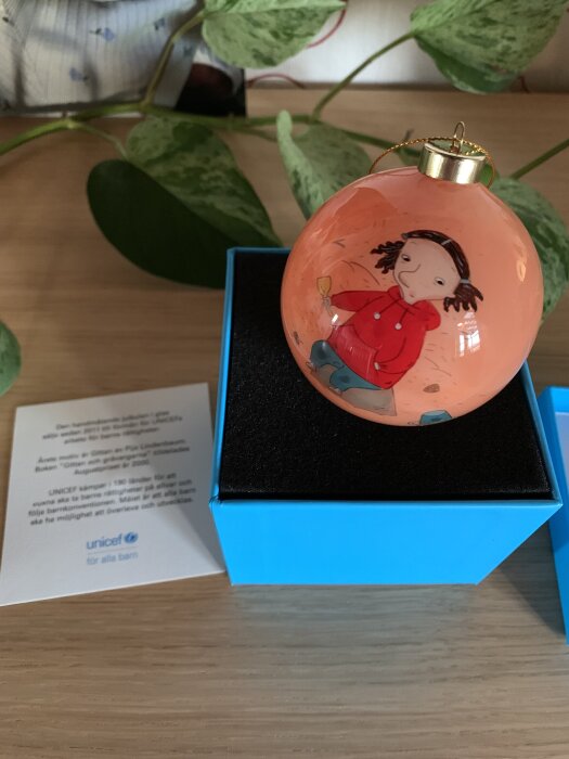 Julgranskula med illustration av barn i röd jacka, liggande i en blå ask. Vintergrön växt i bakgrunden och informativt kort från UNICEF.