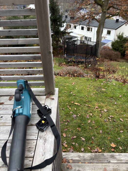 Lövblås ligger på trätrappa med batterier bredvid, omgiven av höstlöv och trädgård med utsikt över hus och studsmatta i bakgrunden.