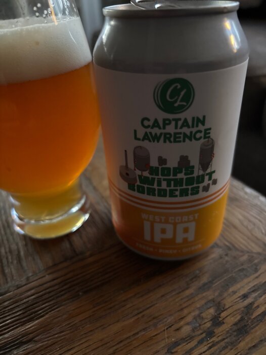 Burk med Captain Lawrence öl "Hops Without Borders" och ett glas öl på ett träbord.