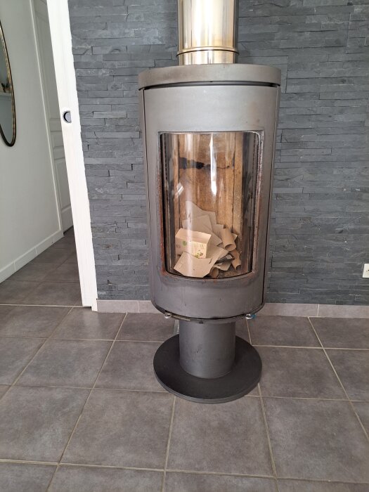 Kamin med glasdörr på mörk stenvägg och grått klinkergolv.