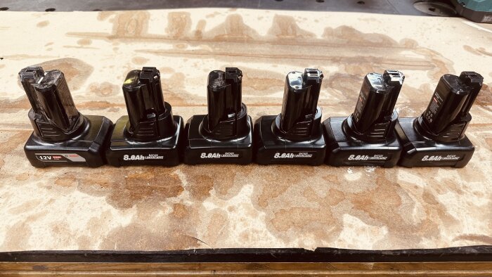 Sex laddningsbara batterier, varav ett Bosch 12V och fem 8.0Ah kopior, står uppradade på ett slitet träbord i en verkstadsmiljö.