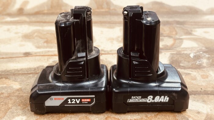 Två Bosch batterier visas sida vid sida, ett 12V 6.0Ah och ett 12V 8.0Ah, på en bakgrund av trä.