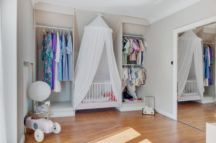 Barnrum med en säng täckt av ett vitt draperi, samt två öppna garderober med vuxen- och barnkläder. På golvet står en leksaksbil och en korglampa.