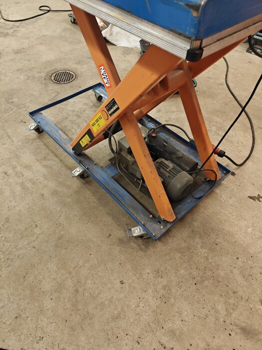 Lyftbord på garagegolv med orange ram och blå plattform, utrustat med länkhjul. Elektrisk motor synlig. Diskussion om stabilitet och enkel flytt.