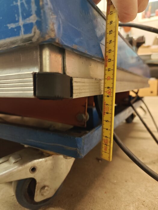 Lyftbord på hjul med måttband visar avstånd till golvet, omgiven av kablar, på garagegolv för stabilitetsförbättring utan länkhjul.