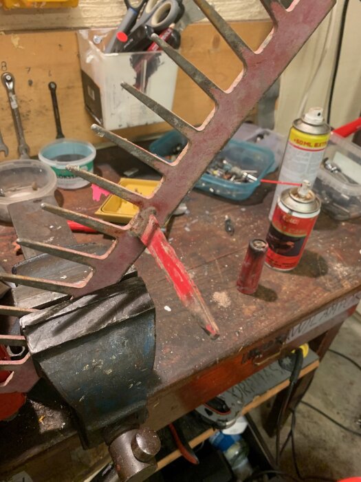 Urfasad kratta utan träskaft fastspänd i ett skruvstäd på en arbetsbänk, omgiven av verktyg och burkar.