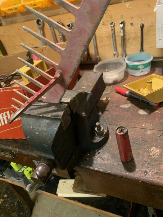 Krattdel i ett skruvstäd på arbetsbänk i verkstad med verktyg i bakgrunden.