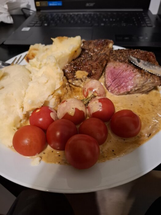 Tallrik med entrecote, potatismos, brunsås och körsbärstomater, framför en laptop på ett skrivbord.