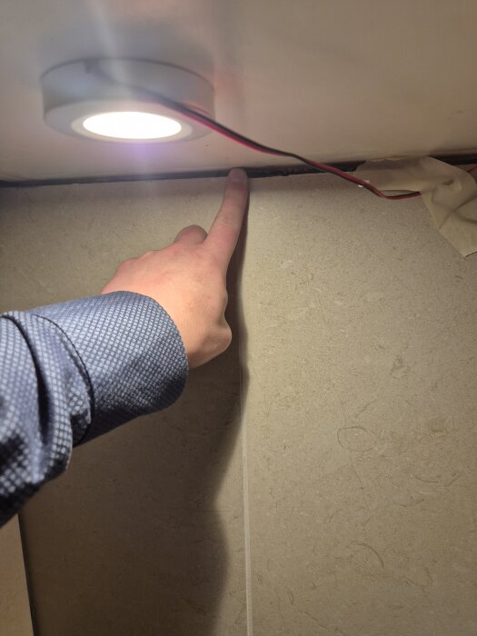 Hand pekar på skarv mellan klinkerplatta och överskåp i köket, belyst av en underliggande lampas sken.