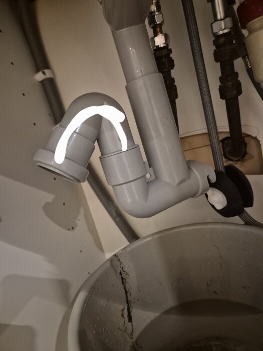Grått avloppsrör under diskbänk med en markerad böjd del, monterat med andra rör och kopplingar, över en hink med smutsigt vatten.