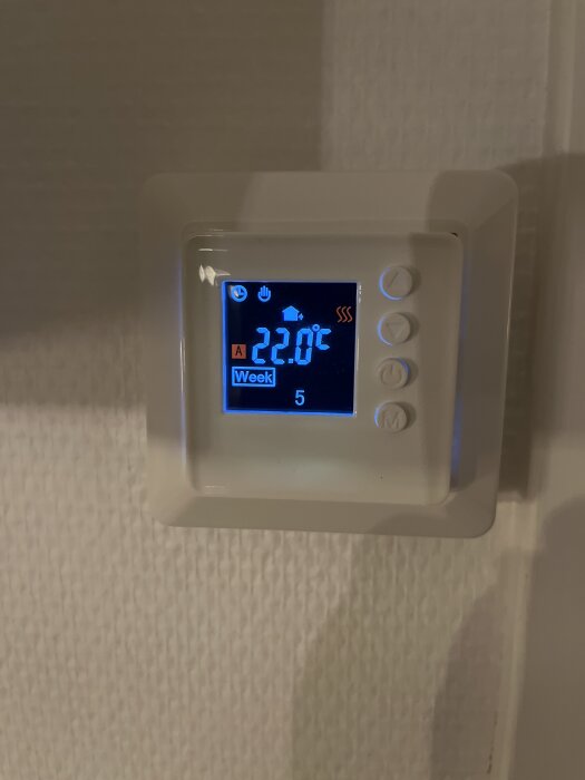 Termostat på en vägg visar 22,0°C med inställningar för vecka 5 och ikoner för värmeindikator och strömläge.