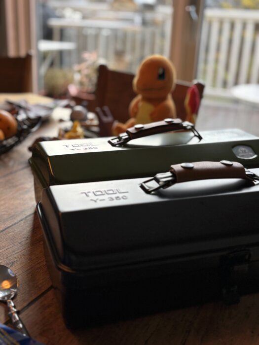 Två verktygslådor på ett bord, en i grönt och en i svart, med en orange leksaksfigur i bakgrunden.