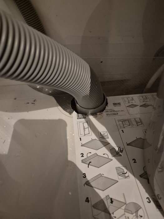 Grå flexibel rörledning kopplad in i en vägg, monteringsanvisningar synliga på ytan under röret.