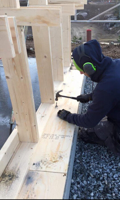 Person som hamrar spik i träregel på byggarbetsplats, iförd hörselskydd och handskar.